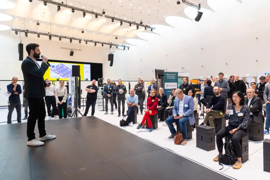 Mann steht auf Bühne. Menschen sitzen vor ihm und hören zu.